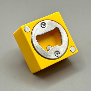 Op de afbeelding: Een heldergele flesopener met een zilveren metalen opener. De opener is vierkant met een cirkelvormige opening en twee schroeven. Twee kleine zilveren magneten bevinden zich aan de zijkanten van de gele vierkant.
