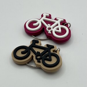 May include: Two bicycle keychain charms. One is pink with a white bicycle and the other is beige with a black bicycle.