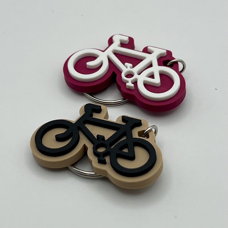 May include: Two bicycle keychain charms. One is pink with a white bicycle and the other is beige with a black bicycle.