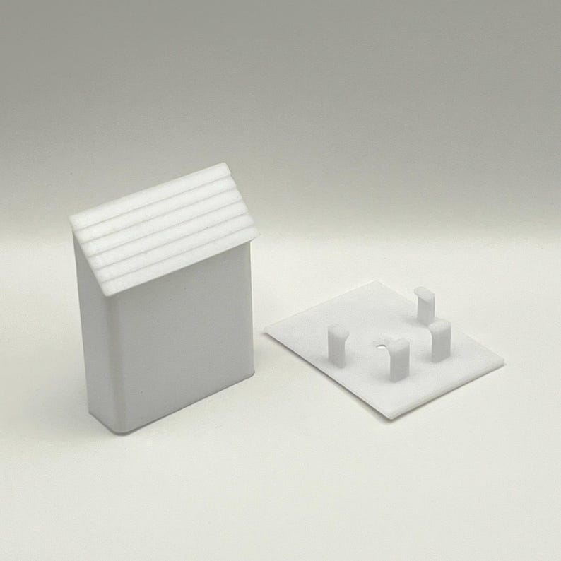 May include: White plastic model of a small building with a gabled roof and a base with four small pillars. The model is designed to be used as a miniature building for a model train set.