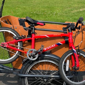 May include: A red Wiggins Macon bicycle with black tires and a black seat. The bicycle is positioned in front of a wooden cargo box. The bicycle has a chain and a gear system. The bicycle has the word "Wiggins" on the frame.