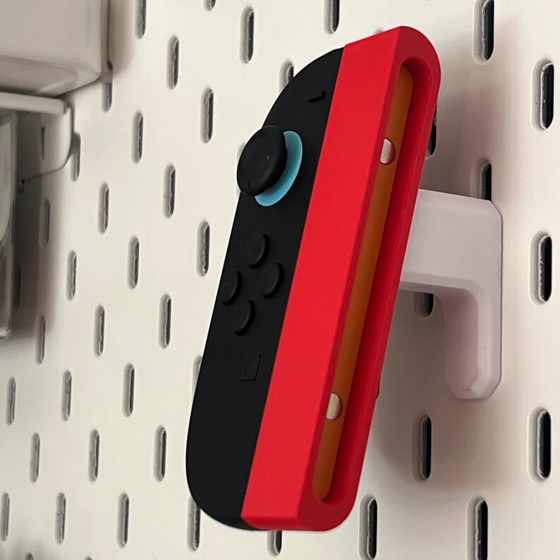 Puede incluir: Un mando Joy-Con de Nintendo Switch rojo y negro est&aacute; montado en una placa perforada blanca con un soporte blanco. El mando tiene botones negros y un joystick azul. La placa perforada tiene ranuras verticales espaciadas.