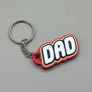 May include: A keychain with a red, yellow, and white design that reads "DAD". The keychain has a silver ring and chain. The keychain is a perfect gift for Father's Day or any special occasion.