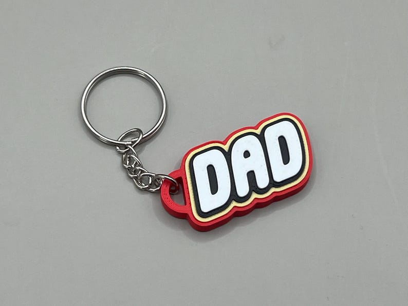 May include: A keychain with a red, yellow, and white design that reads "DAD". The keychain has a silver ring and chain. The keychain is a perfect gift for Father's Day or any special occasion.