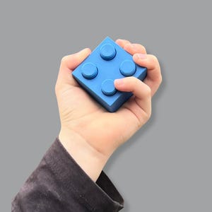 May include: A bright blue building block with four circular studs on top. The block is held in a hand against a gray background. The block is a solid, vibrant color.