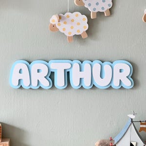 May include: A light blue wooden name sign with the name "ARTHUR" in white letters. The letters are outlined in blue and have a rounded, bubbly shape. The sign is hanging on a light blue wall.