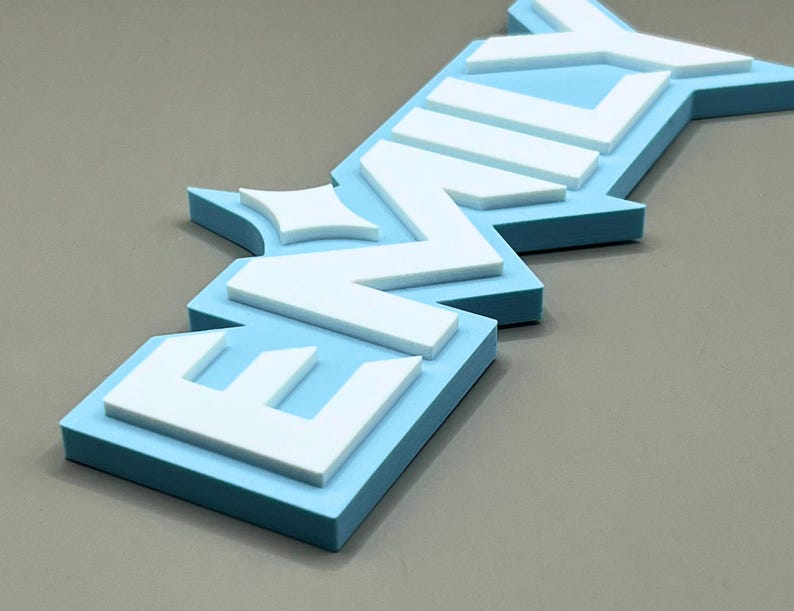 May include: A 3D sign spelling out the word "ENVY" in a layered design. The base layer is a light blue, with white letters stacked on top. The sign is angled, with the letters appearing to be slightly raised.