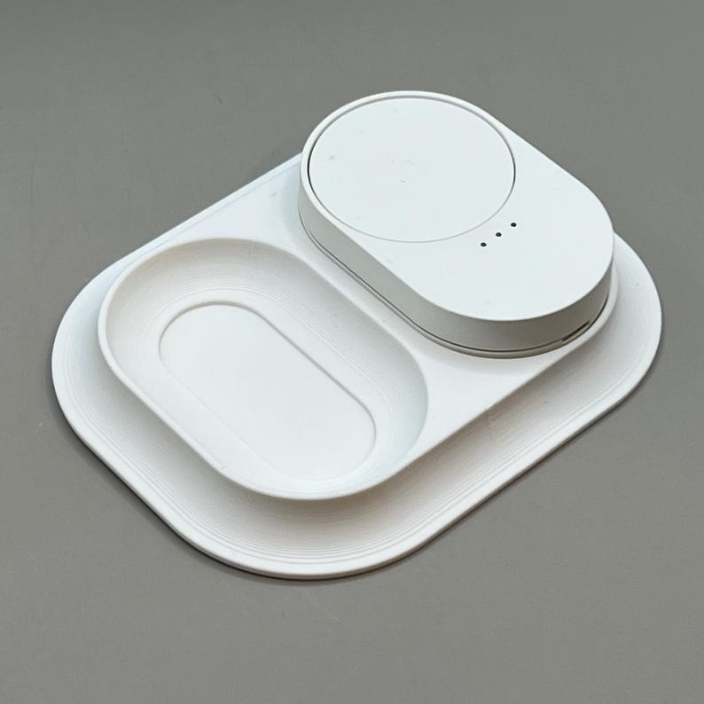 Pu&ograve; includere: Una stazione di ricarica bianca per pi&ugrave; dispositivi con base rettangolare arrotondata. Presenta un pad di ricarica circolare e un vassoio di forma ovale. Tre piccole spie luminose sono visibili sull'unit&agrave; di ricarica. Design minimalista e moderno.