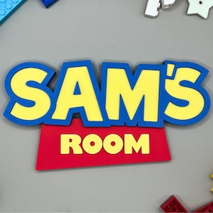 May include: A colorful sign that reads "SAM'S ROOM" in yellow letters with a blue outline, set against a red background. The sign is surrounded by toy building blocks in various colors, including blue, yellow, and red, on a gray surface.