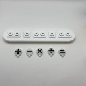 May include: A white plastic tray with six round holes and five white plastic buttons with black mathematical symbols: plus, minus, multiply, divide, and equals.