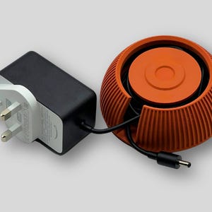 Puede incluir: Un adaptador de corriente negro con un enchufe de tres clavijas, conectado a un enrollador de cable naranja. El enrollador tiene un dise&ntilde;o redondo y acanalado con una parte superior circular. El cable es negro y conecta el adaptador al enrollador.