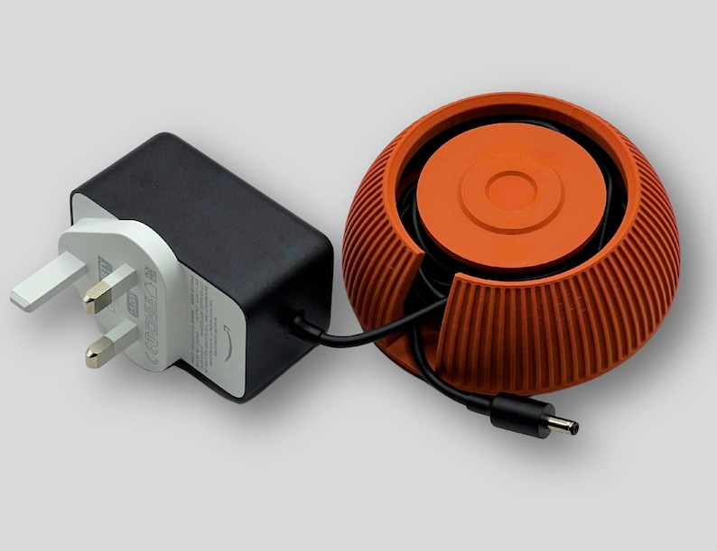 Puede incluir: Un adaptador de corriente negro con un enchufe de tres clavijas, conectado a un enrollador de cable naranja. El enrollador tiene un dise&ntilde;o redondo y acanalado con una parte superior circular. El cable es negro y conecta el adaptador al enrollador.
