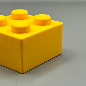 Op de afbeelding: Een heldergele bouwsteen met vier verhoogde ronde noppen aan de bovenkant. De blok is een solide, rechthoekige vorm met een getextureerd oppervlak, perfect voor creatief bouwen en spelen.