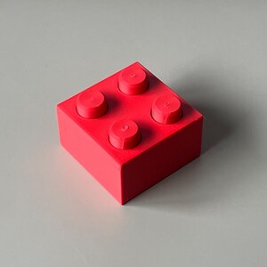 May include: A bright red building block with four circular studs on top. The block is a solid, square shape, and the studs are evenly spaced. The block is set against a neutral gray background.