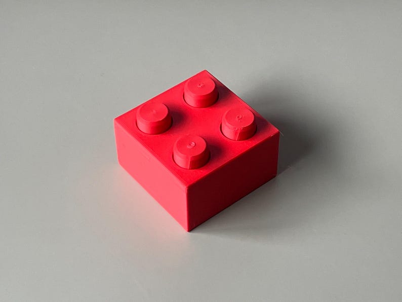 May include: A bright red building block with four circular studs on top. The block is a solid, square shape, and the studs are evenly spaced. The block is set against a neutral gray background.