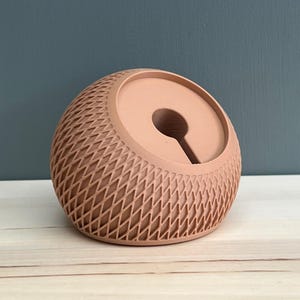 May include: A peach-colored, spherical desk accessory featuring a textured, diamond-patterned lattice exterior and a smooth, circular recessed center with a vertical slot opening, designed to hold small items or cables securely.