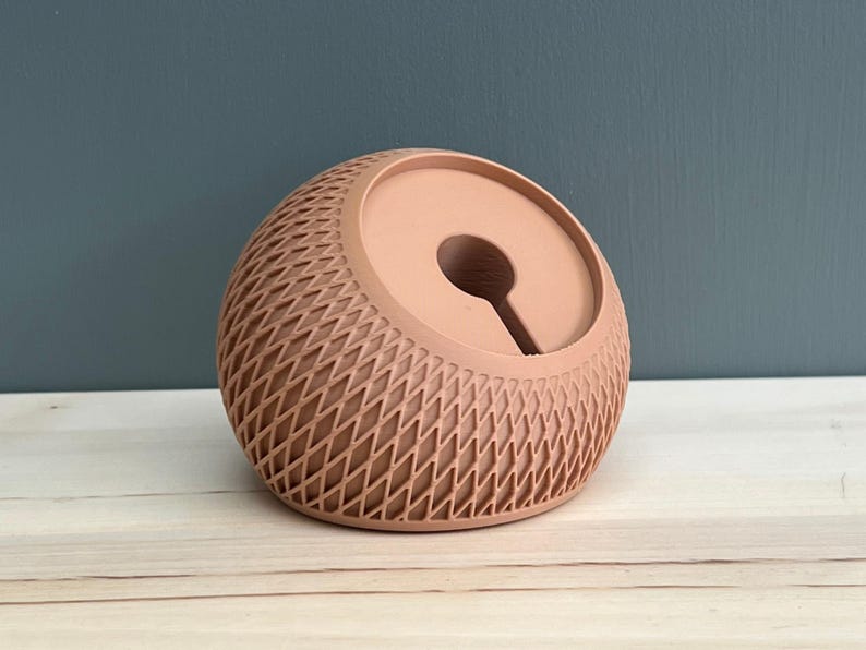 May include: A peach-colored, spherical desk accessory featuring a textured, diamond-patterned lattice exterior and a smooth, circular recessed center with a vertical slot opening, designed to hold small items or cables securely.