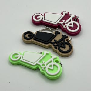 May include: Three colorful keychains shaped like cargo bikes with white, black, and green accents. The keychains are made of a rubbery material and have a metal ring for attaching keys.