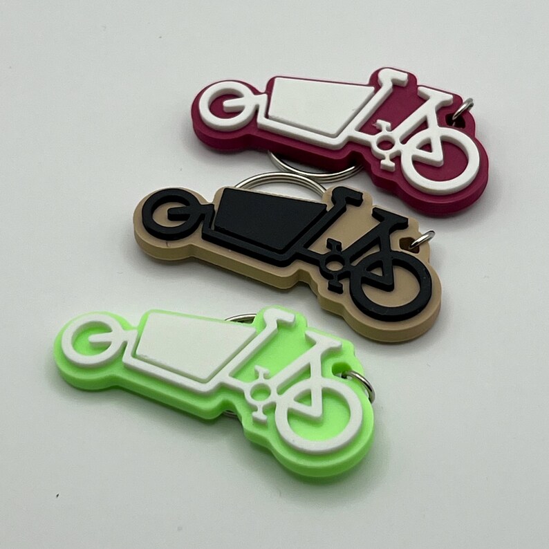 May include: Three colorful keychains shaped like cargo bikes with white, black, and green accents. The keychains are made of a rubbery material and have a metal ring for attaching keys.