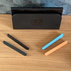 May include: Black Nintendo Switch dock with the Switch console inserted. Four colorful Joy-Con rail covers are arranged in front of the dock. Two are black with orange and blue accents, and two are blue and orange.
