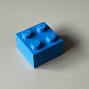 May include: A bright blue building block with four raised studs on top. The block is square and has a smooth, solid appearance. The image is taken from a slightly elevated angle, showcasing the block's form.