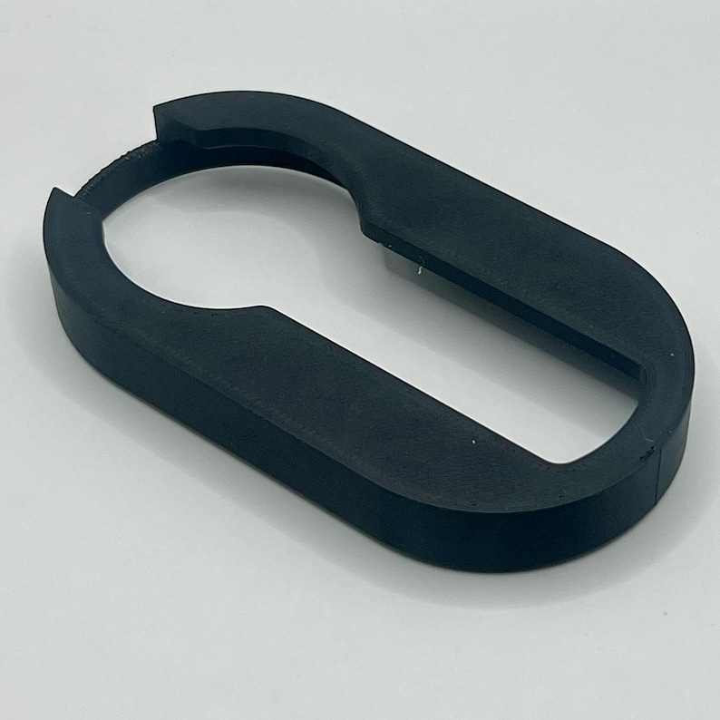 May include: A black, oval-shaped tool with a curved handle and a keyhole-like opening. The tool is made of a matte material and appears to be 3D-printed. The design suggests it may be used for gripping or opening something.