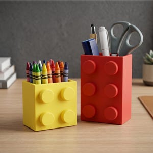 May include: A yellow square pencil holder and a taller red rectangular pencil holder, both designed to resemble building blocks with circular studs on the front. The yellow holder contains various colourful crayons, while the red holder holds a blue stapler, a white pen, and grey scissors.