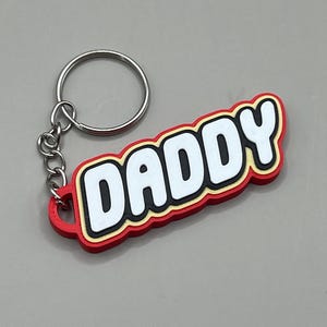 May include: A red keychain with the word "DADDY" in white, outlined in black and yellow. The keychain has a silver metal ring and chain.