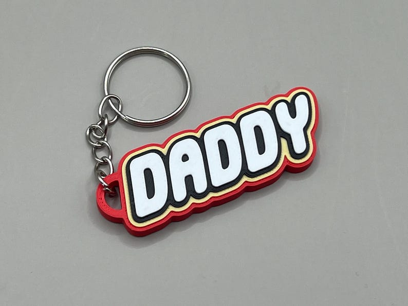 May include: A red keychain with the word "DADDY" in white, outlined in black and yellow. The keychain has a silver metal ring and chain.