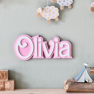 Puede incluir: Un letrero de nombre de madera rosa con el nombre "Olivia" en letras blancas. El letrero tiene un dise&ntilde;o de espiral decorativo alrededor de la primera letra.