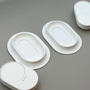 Pu&ograve; includere: Vassoi bianchi di forma ovale e piccoli dispositivi arrotondati sono disposti su una superficie grigia. I vassoi hanno un centro ovale rialzato all'interno di una base ovale pi&ugrave; grande. I dispositivi hanno un design liscio e arrotondato.