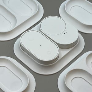 Pu&ograve; includere: Stazione di ricarica bianca stampata in 3D con due basi di ricarica ovali e una base circolare. Diversi vassoi bianchi con ritagli ovali circondano la stazione di ricarica. Design minimalista e moderno.