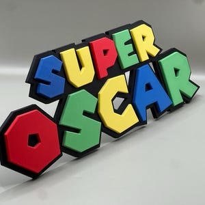 Puede incluir: Un letrero colorido que dice "SUPER OSCAR" en letras de bloque en negrita. Las letras son rojas, amarillas, verdes y azules, con contornos negros. El letrero es probablemente un art&iacute;culo decorativo o un regalo personalizado.