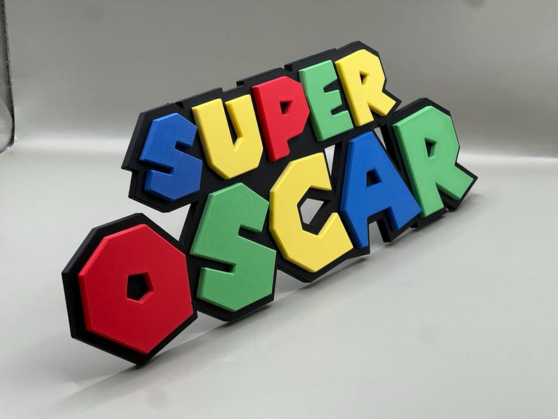Puede incluir: Un letrero colorido que dice "SUPER OSCAR" en letras de bloque en negrita. Las letras son rojas, amarillas, verdes y azules, con contornos negros. El letrero es probablemente un art&iacute;culo decorativo o un regalo personalizado.