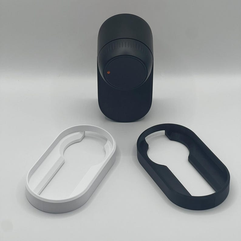 May include: A black electronic device with a circular dial and a red indicator light, accompanied by a white and a black oval-shaped accessory. The items are displayed against a plain background, suggesting a tech or gadget product.