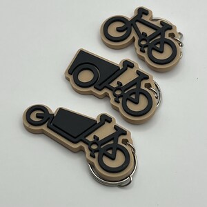 May include: Three keychains featuring a black silhouette of a bicycle with a trailer attached. The keychains are made of a beige rubber-like material.