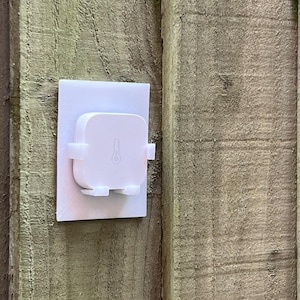 May include: A white square-shaped device with a temperature sensor mounted on a white plastic wall plate. The device is attached to a wooden wall.