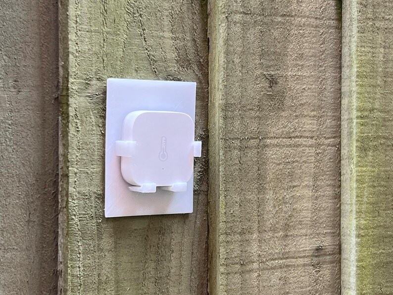 May include: A white square-shaped device with a temperature sensor mounted on a white plastic wall plate. The device is attached to a wooden wall.