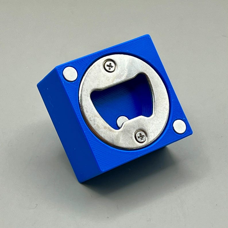 Op de afbeelding: Een blauwe vierkante flesopener met een zilveren metalen opener en twee schroeven. Twee witte cirkelvormige magneten bevinden zich aan de zijkanten van de blauwe vierkant. Een handig baraccessoire.