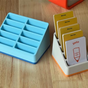 May include: A light blue plastic storage rack with ten slots and a white plastic storage rack with five slots containing four yellow cards labelled Stories and one white card with the red text yoto and a drawing of a smiling pencil.