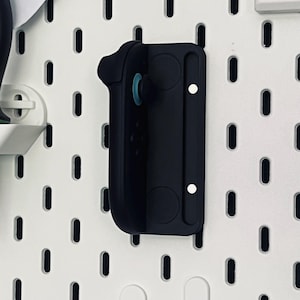 Peut inclure: Un support mural noir pour manette de jeu, en plastique. Il pr&eacute;sente un design incurv&eacute; pour maintenir la manette et est mont&eacute; sur une plaque perfor&eacute;e blanche avec des trous ovales. Le support a deux aimants circulaires et une vis blanche.
