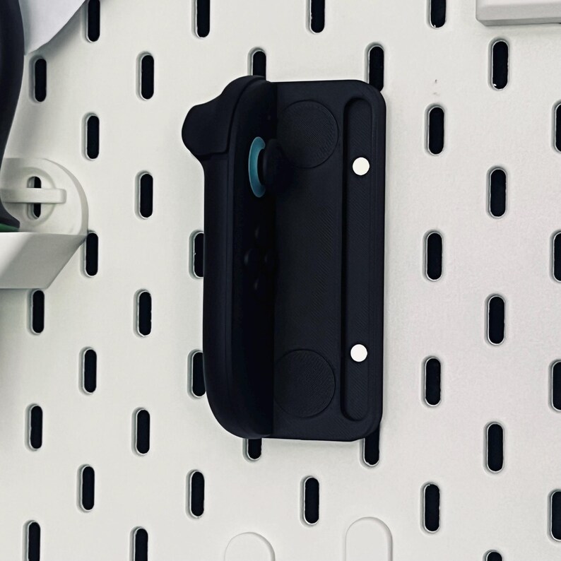 Peut inclure: Un support mural noir pour manette de jeu, en plastique. Il pr&eacute;sente un design incurv&eacute; pour maintenir la manette et est mont&eacute; sur une plaque perfor&eacute;e blanche avec des trous ovales. Le support a deux aimants circulaires et une vis blanche.