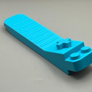 May include: A bright turquoise game controller-shaped object. The controller has a cross-shaped button and two round buttons. The top surface has a series of parallel grooves. The object is made of a solid, smooth material.