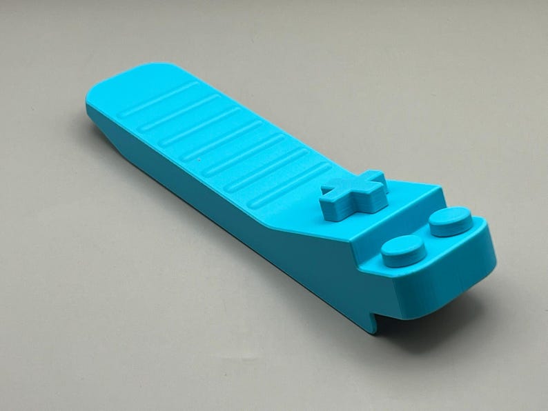 May include: A bright turquoise game controller-shaped object. The controller has a cross-shaped button and two round buttons. The top surface has a series of parallel grooves. The object is made of a solid, smooth material.