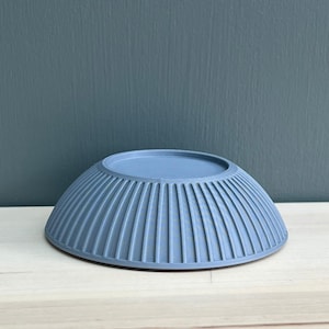 May include: A matte, dusty blue bowl with a rounded shape and a series of vertical, evenly spaced ridges running along the exterior surface. The bowl is shown upside down, revealing a flat, circular base at the top and a wide, flared rim.