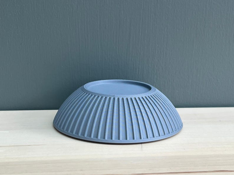 May include: A matte, dusty blue bowl with a rounded shape and a series of vertical, evenly spaced ridges running along the exterior surface. The bowl is shown upside down, revealing a flat, circular base at the top and a wide, flared rim.