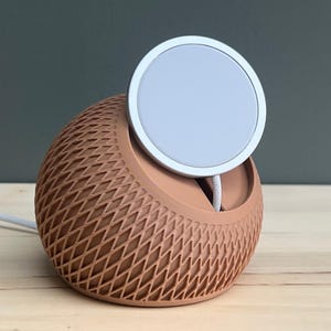 May include: A spherical charging dock with a terracotta-colored lattice exterior and a flat, circular white magnetic charging pad positioned at the top. A white power cable extends from the base of the device, which features a textured, cross-hatched pattern.