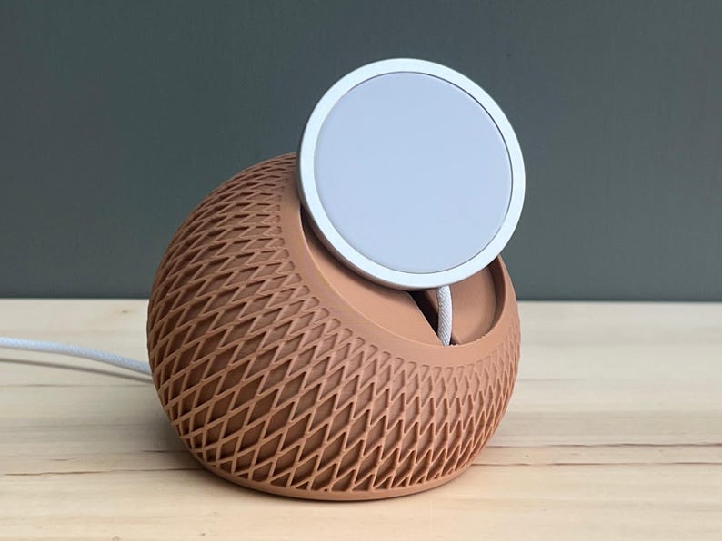 May include: A spherical charging dock with a terracotta-colored lattice exterior and a flat, circular white magnetic charging pad positioned at the top. A white power cable extends from the base of the device, which features a textured, cross-hatched pattern.