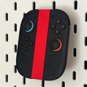May include: Black games controller with a central red stripe. The controller features blue, orange, and white buttons. It is mounted on a white pegboard with evenly spaced holes.