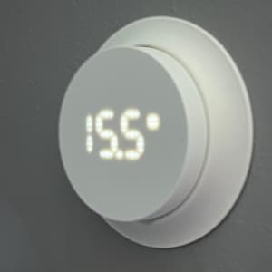 May include: A white, circular thermostat with a digital display showing "15.5&deg;". The thermostat is mounted on a gray wall. The design is modern and minimalist, with a focus on functionality and ease of use.